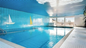 An indoor swimming pool with a tiled wall featuring a sailboat design, a clear ceiling, and a view of the outdoors through large windows. - Vacation apartment Bergkristall with Watzmann view (Berchtesgaden)