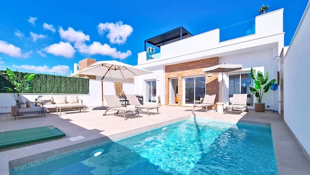 Uma área moderna de piscina ao ar livre, com espreguiçadeiras e guarda-sóis, rodeada por um edifício branco com grandes janelas e um deck de madeira.