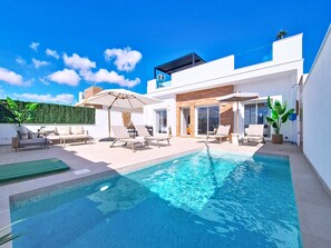 A modern outdoor pool area with lounge chairs and umbrellas, surrounded by a white building with large windows and a wooden deck. - Villa with private pool and roof terrace (San Javier)