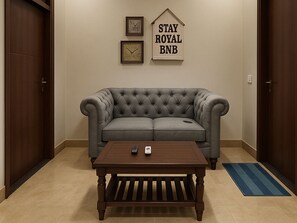 A room with a grey tufted sofa, a wooden coffee table, two framed clocks, and a sign that reads 'STAY ROYAL BNB'. - Stay Royal Villa BNB GF (Kharar)