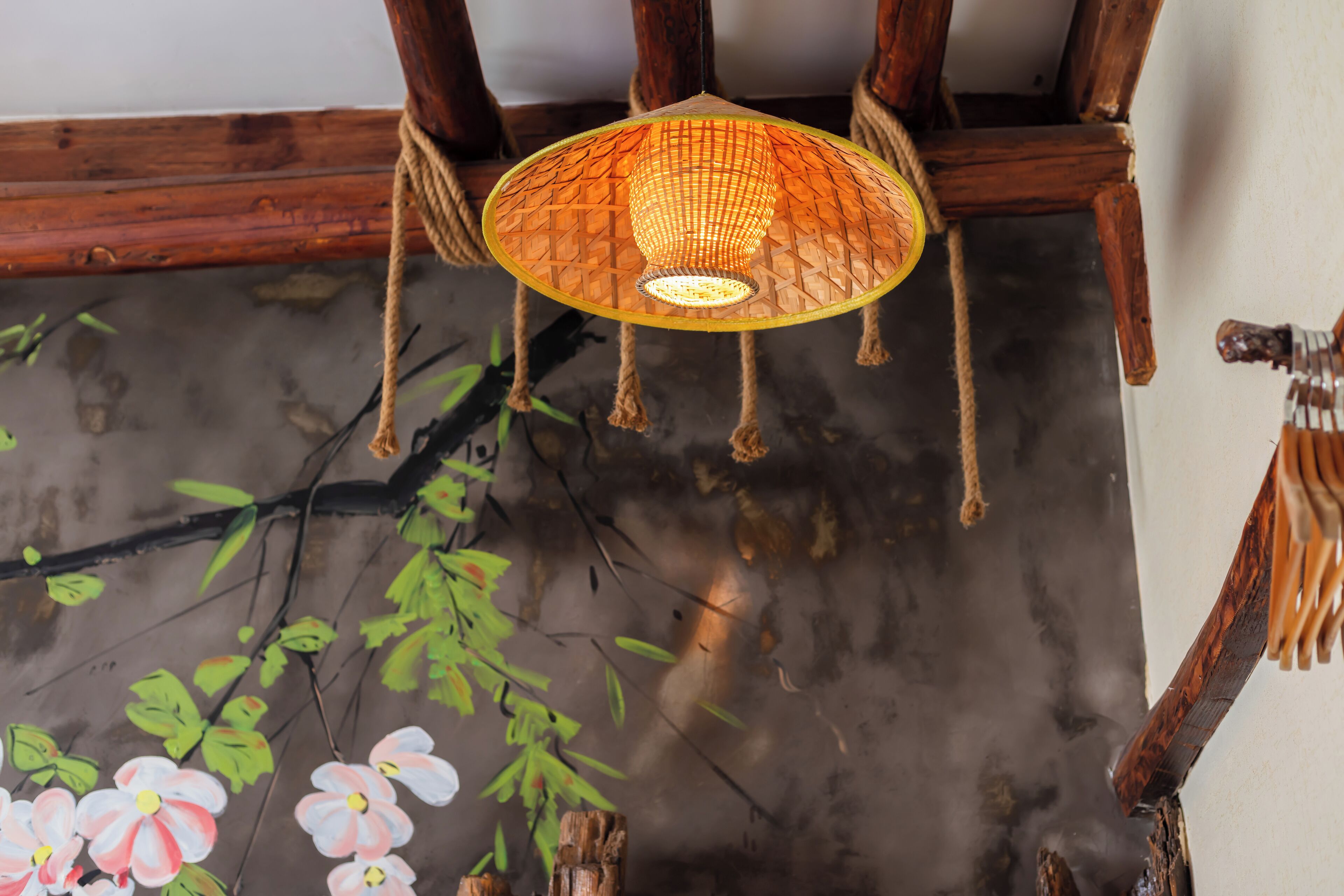 Una lámpara colgante tradicional con pantalla trenzada, patas de silla de madera y una rama pintada con flores.