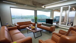 A modern living room with leather sofas, a coffee table, and a flat-screen TV. Large windows offer a view of the outdoors. - Amber Villa By Spandan Holiday Homes (Trimbak)