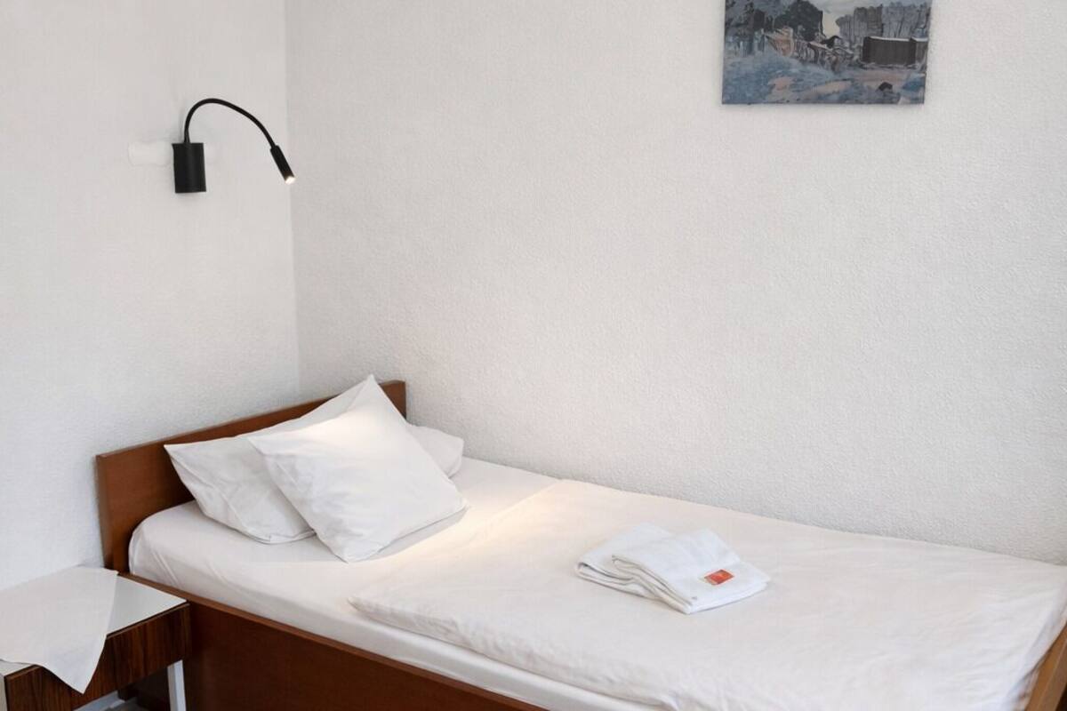 A single bed with a wooden frame, white bedding, and a small bedside table. A wall-mounted lamp is visible on the left side of the bed.
