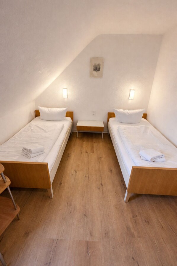 A small, triangular room with two single beds, each with a wooden frame and white bedding. There are two bedside tables with lamps, and the room has a wooden floor. - Hotel Haus Schwan (Cologne)