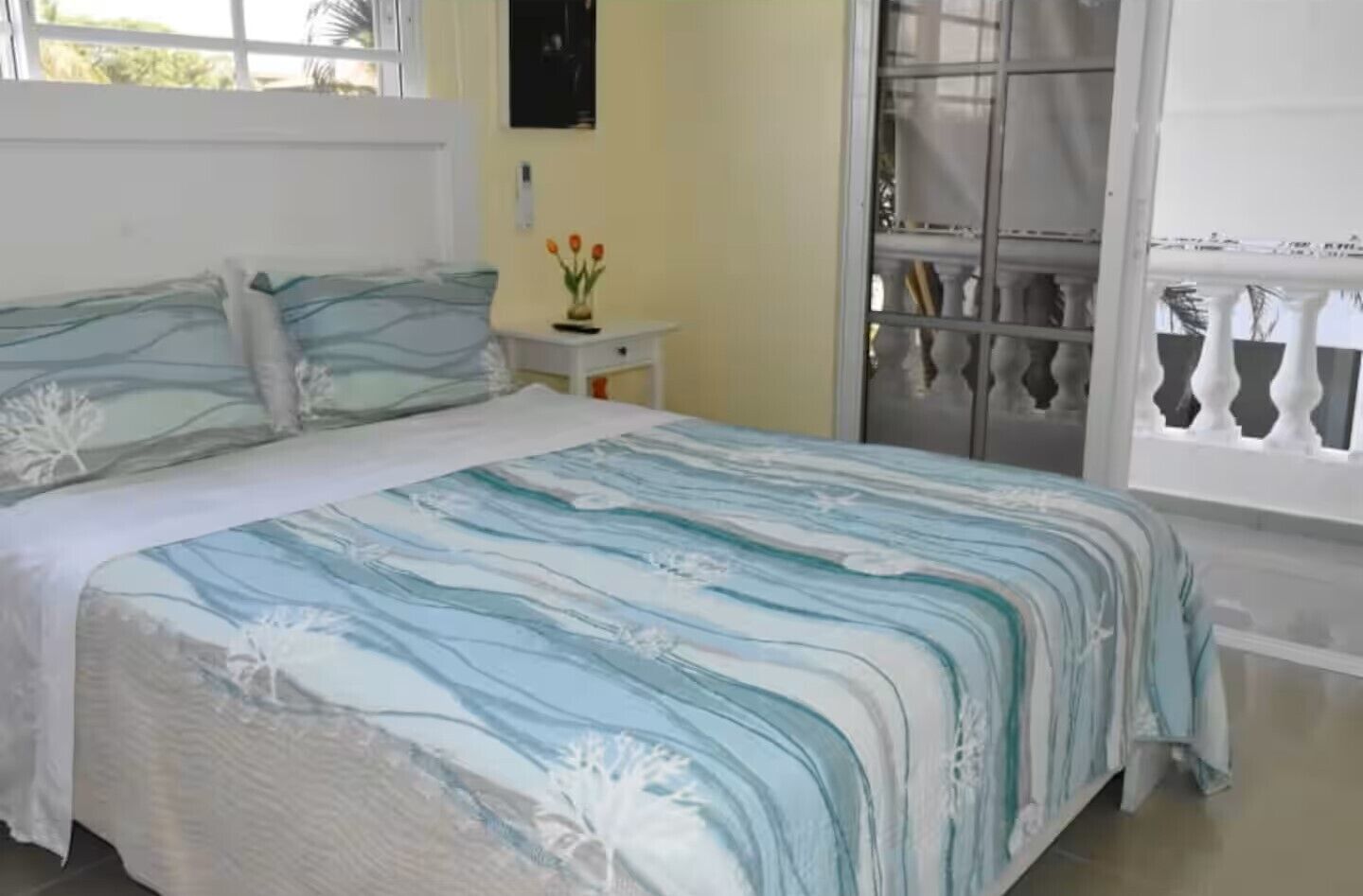 Un dormitorio con cama, mesita de noche, ventana con vista a un balcón y un reloj en la pared.