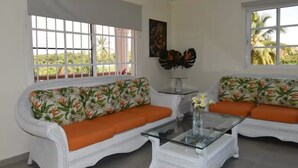 A room with two white wicker sofas with orange cushions, a glass coffee table, and large windows with a view of greenery. - Punta Cana Beach Luxury apartment 2 bedroom 4 minute walk to beach (Punta Cana)