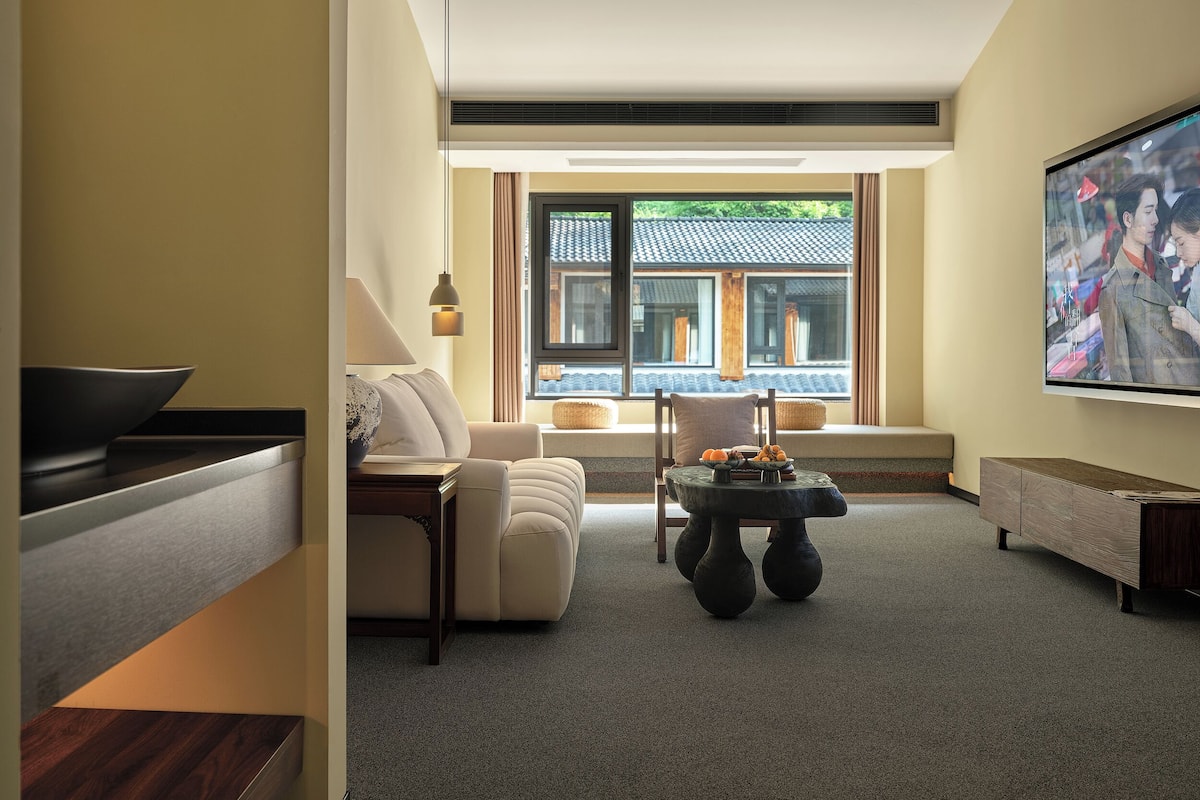 Un soggiorno moderno con un divano, un tavolino da caffè con della frutta, una TV e vista sull'architettura tradizionale attraverso ampie finestre.
