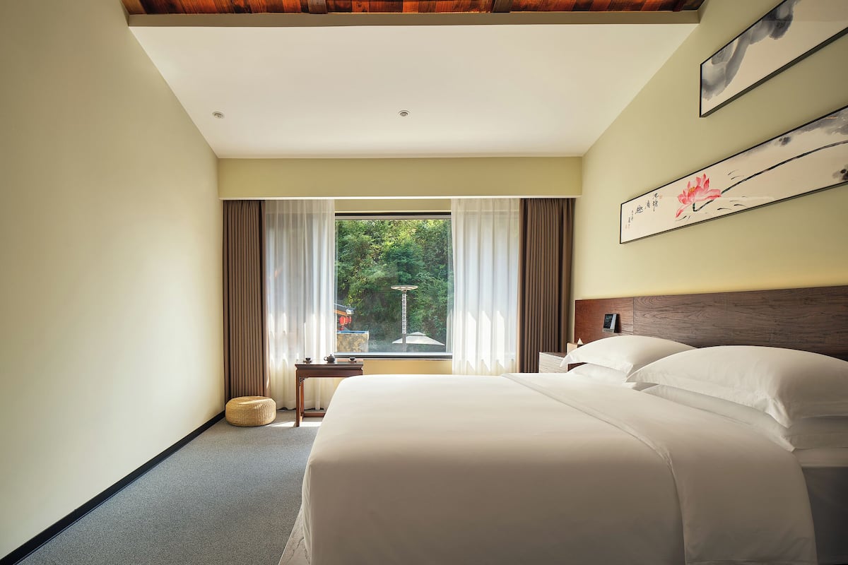 Una camera d'albergo con un letto grande, un soffitto in legno, vista su del verde all'esterno e un piccolo tavolino con una lampada.