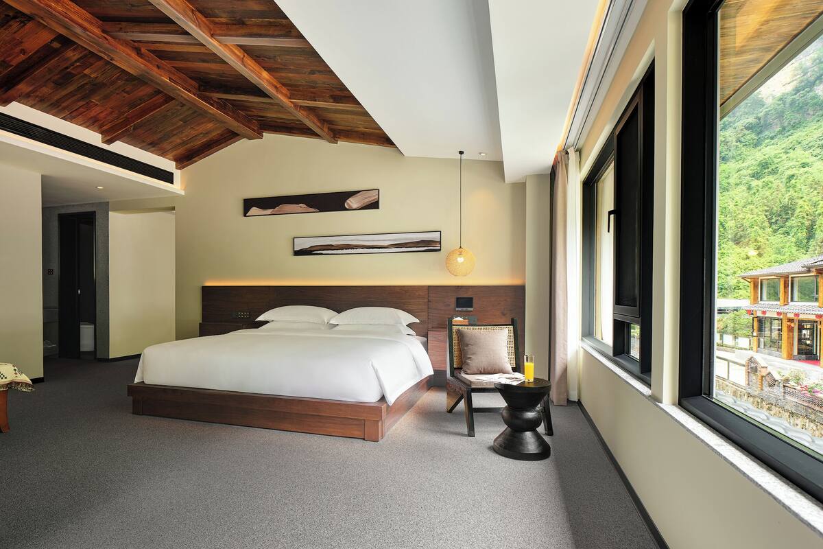 Camera d'albergo moderna con un letto grande, soffitto in legno e vista su un edificio tradizionale attraverso un'ampia finestra.