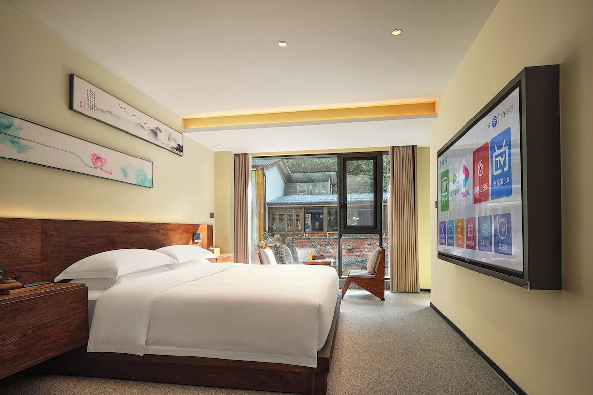 Una camera d'albergo con un letto grande, una testiera in legno, una scrivania con una sedia e un'ampia finestra vista sull'architettura tradizionale.