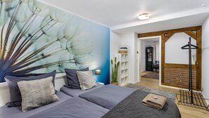 Een slaapkamer met een groot wanddecor, een bed met grijze beddengoed, een nachtkastje met een lamp, en een uitzicht op een aangrenzende kamer met een houten balk en een hangroede.