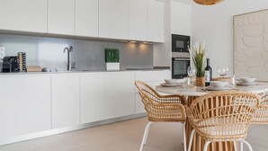 A modern kitchen with white cabinets, a built-in oven, and a dining area with wicker chairs and a round table. - Casa Oceana | Pool - views - gym (Estepona)
