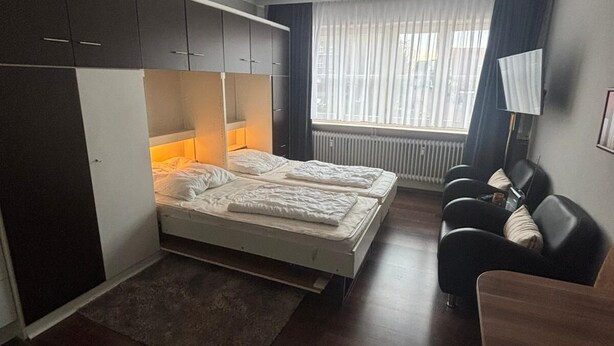 Ein kleines Zimmer mit Bett, Schreibtisch und Stuhl. Es gibt ein Fenster mit Vorhängen, eine Heizung und eine Deckenleuchte.