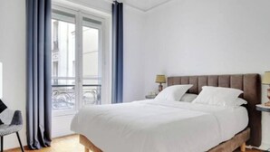 Quarto com uma cama grande, mesa de cabeceira, cadeira e janela com cortinas.