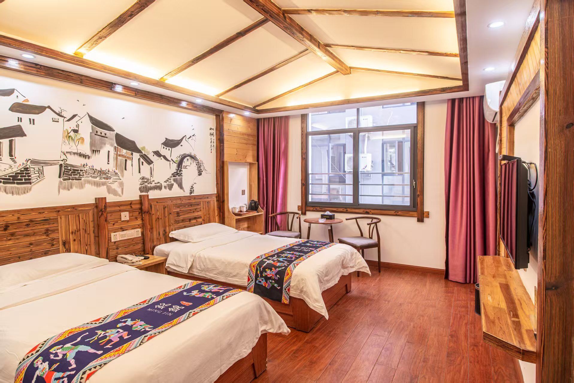 Een hotelkamer met twee bedden, houten hoofdeinden, een muurschildering, een raam met gordijnen en een kleine tafel met stoelen.
