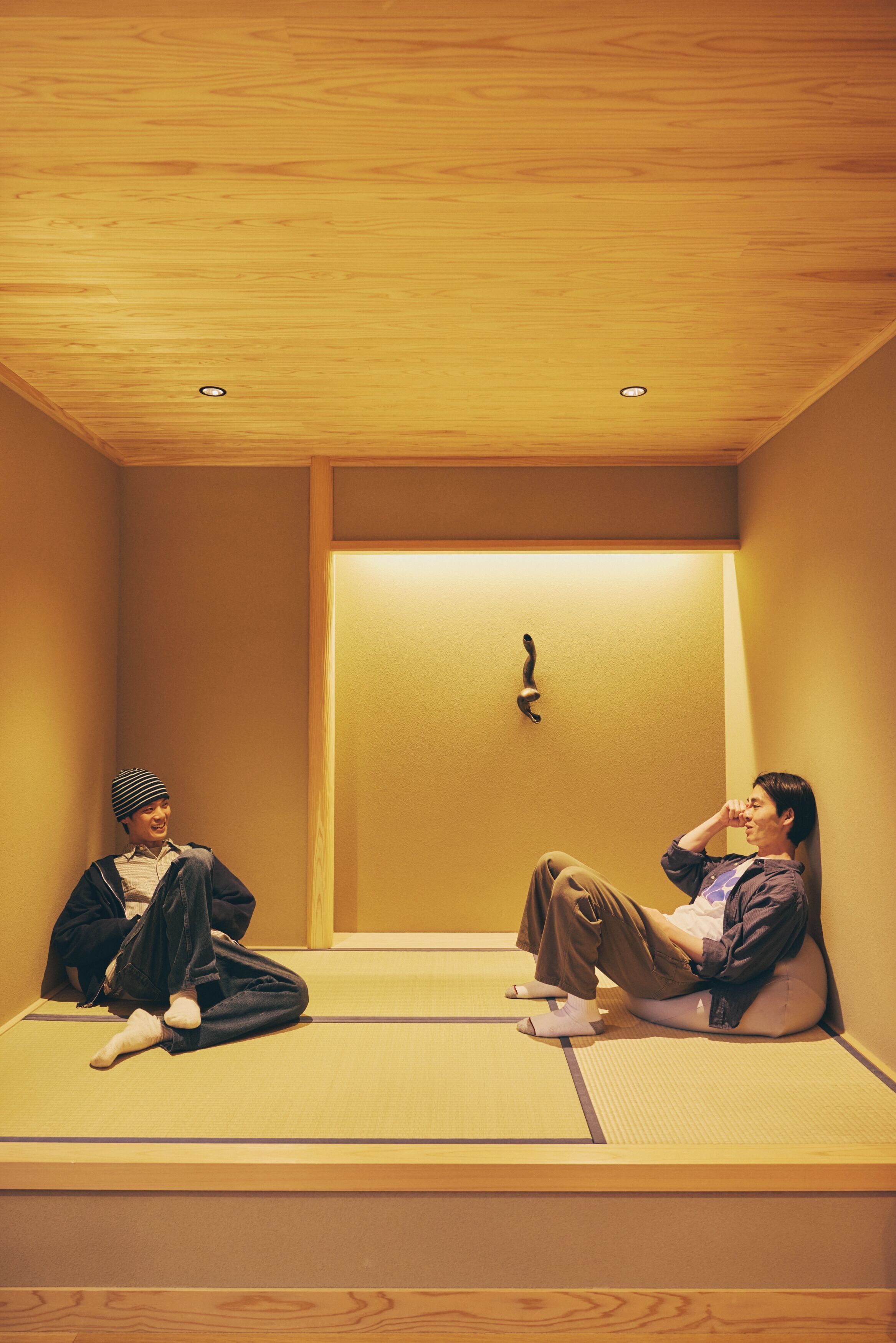 Two individuals sitting on tatami mats in a room with a wooden ceiling and a door with a handle.