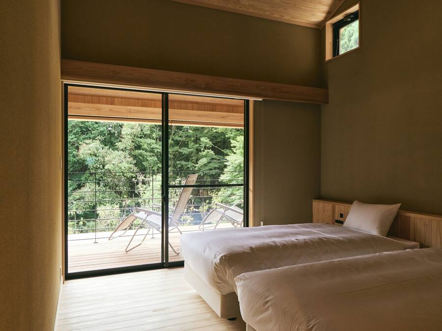 Un dormitorio con un gran ventanal corredera que da a una terraza exterior, suelo de madera y una cama con ropa blanca.
