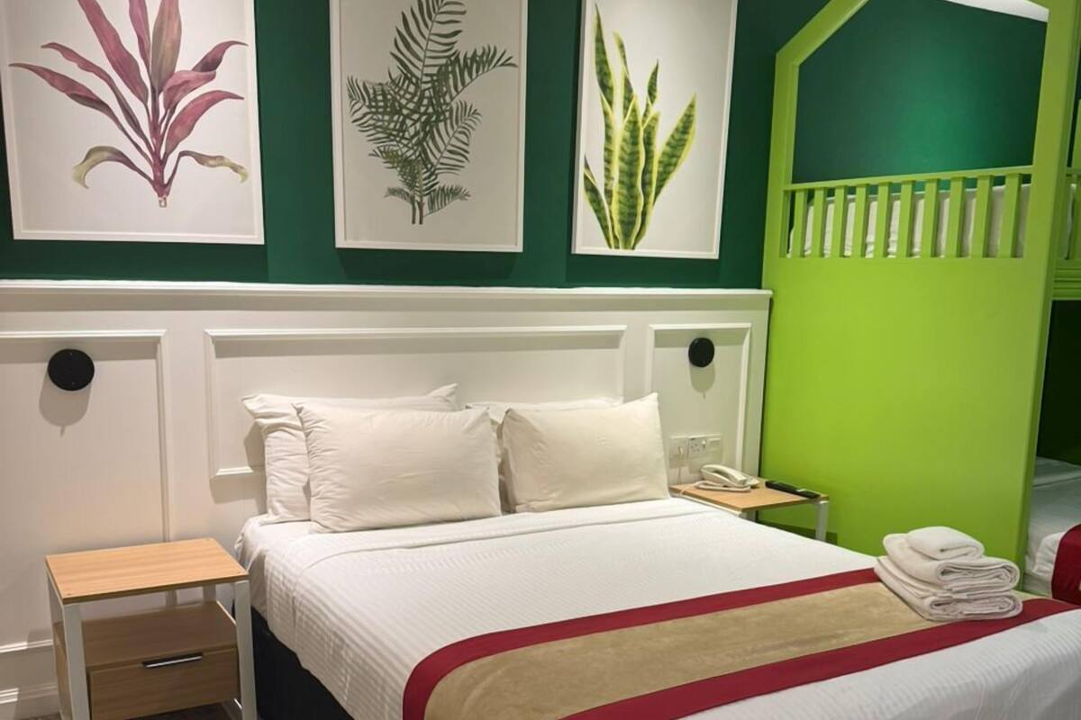 A neatly made bed with white linens and a red accent, a bedside table with a drawer, and a green accent wall with framed botanical prints.