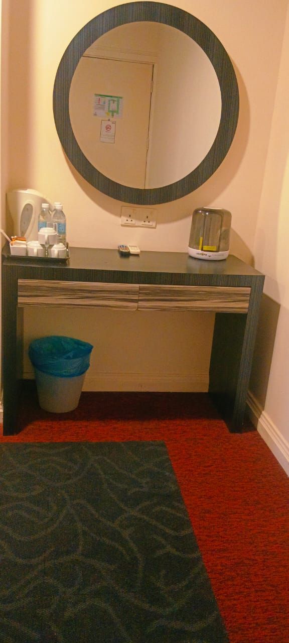 A hotel room with a round mirror above a beige vanity, a blue trash bin, and a red carpet.