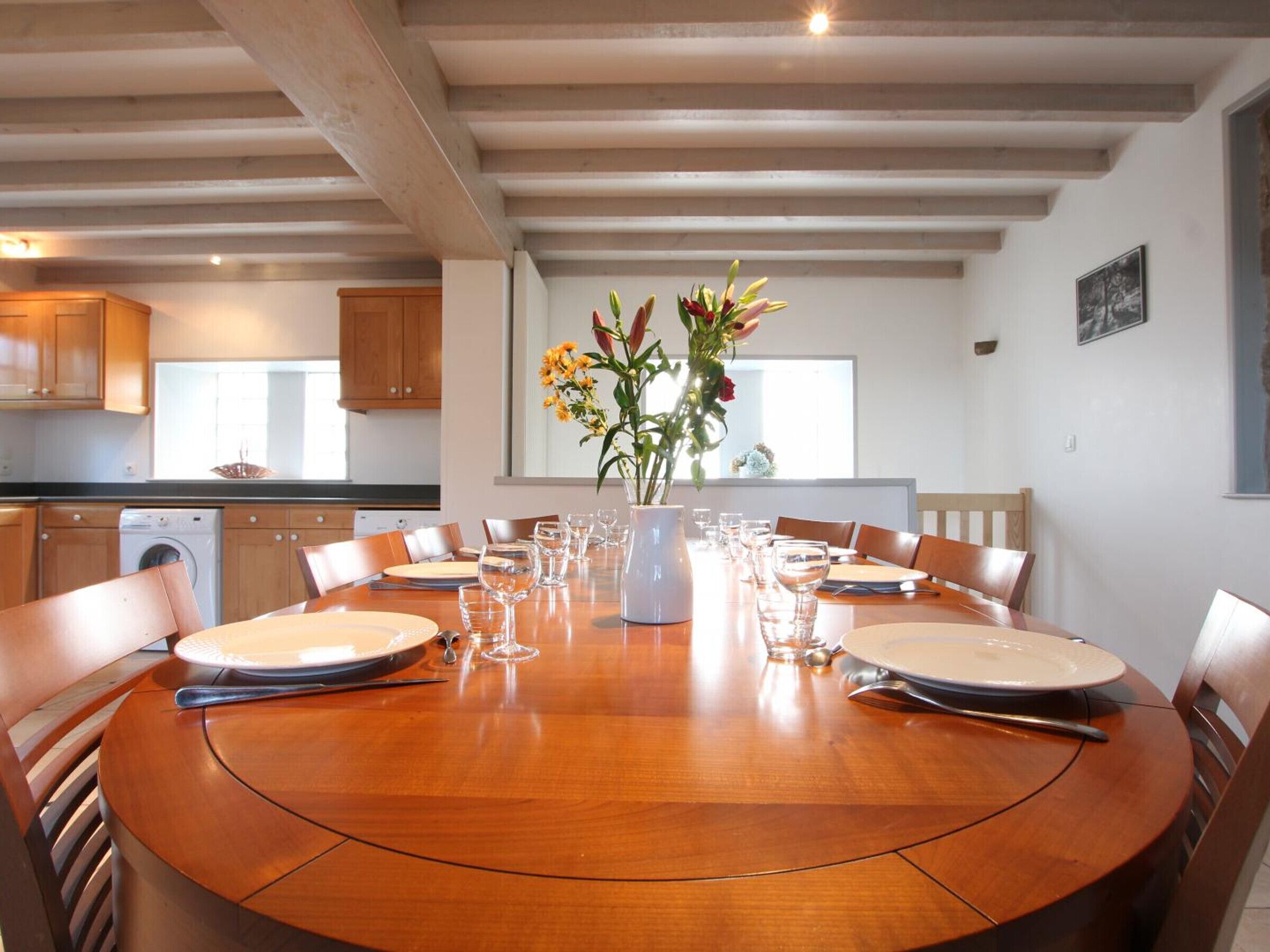 Een eetruimte met een ronde houten tafel voor zes personen, omringd door houten stoelen. In het midden staat een witte vaas met bloemen.