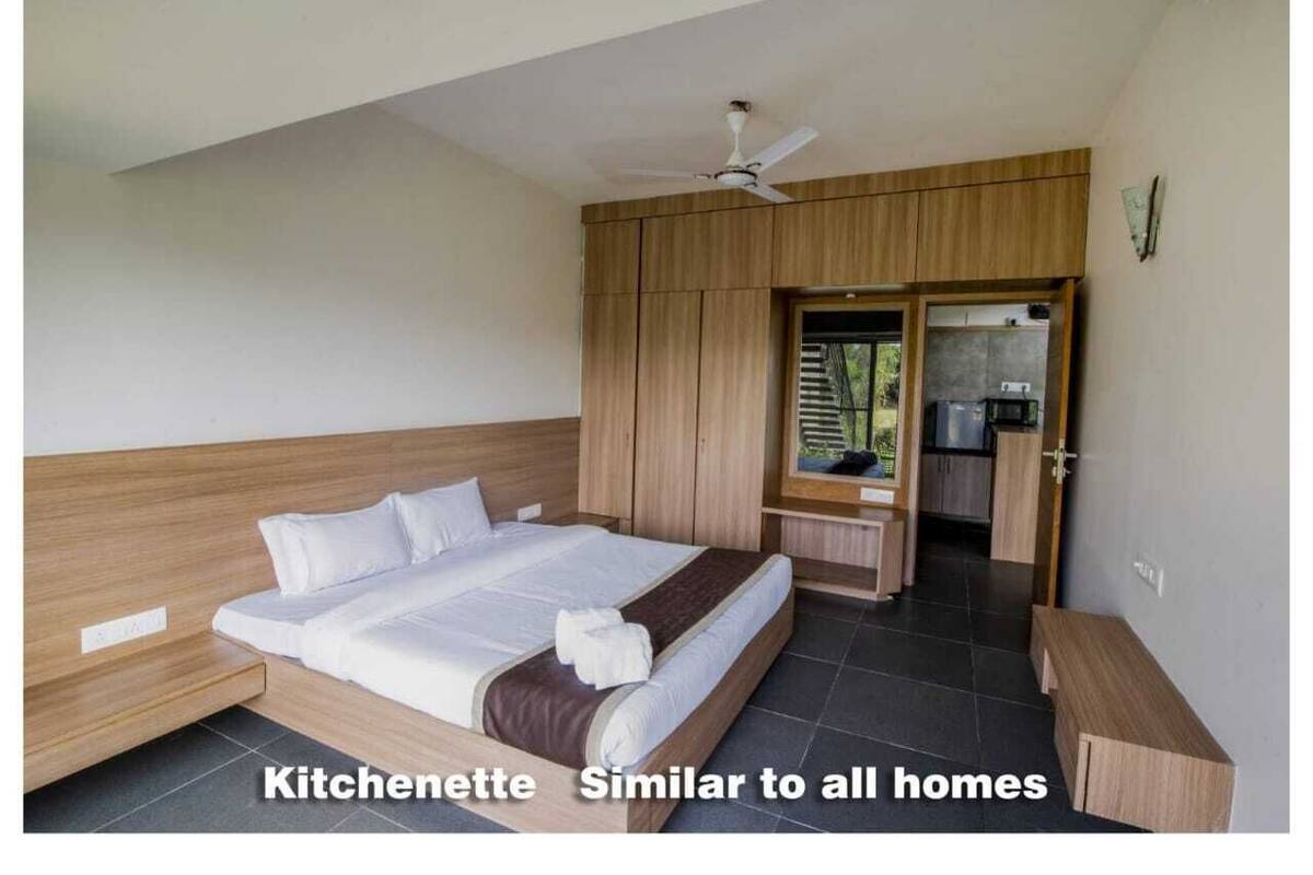 Un dormitorio con cama, cabecera de madera, ventilador de techo, kitchenette y baño visible a través de una puerta abierta.