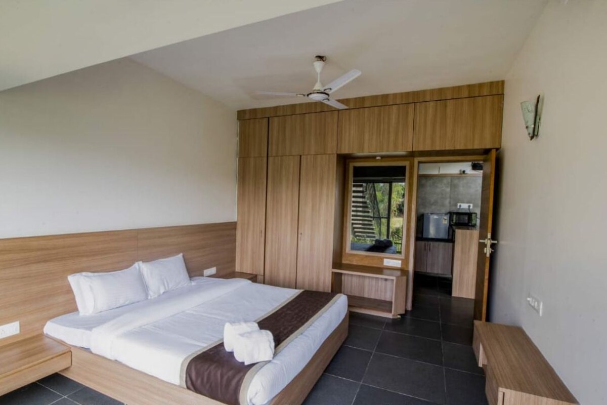 Une chambre à coucher avec un lit, une tête de lit en bois, un ventilateur de plafond, un coin cuisine et une salle de bain visible à travers une porte ouverte.