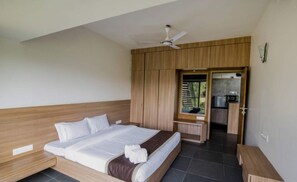 Une chambre à coucher avec un lit, une tête de lit en bois, un ventilateur de plafond, un coin cuisine et une salle de bain visible à travers une porte ouverte.