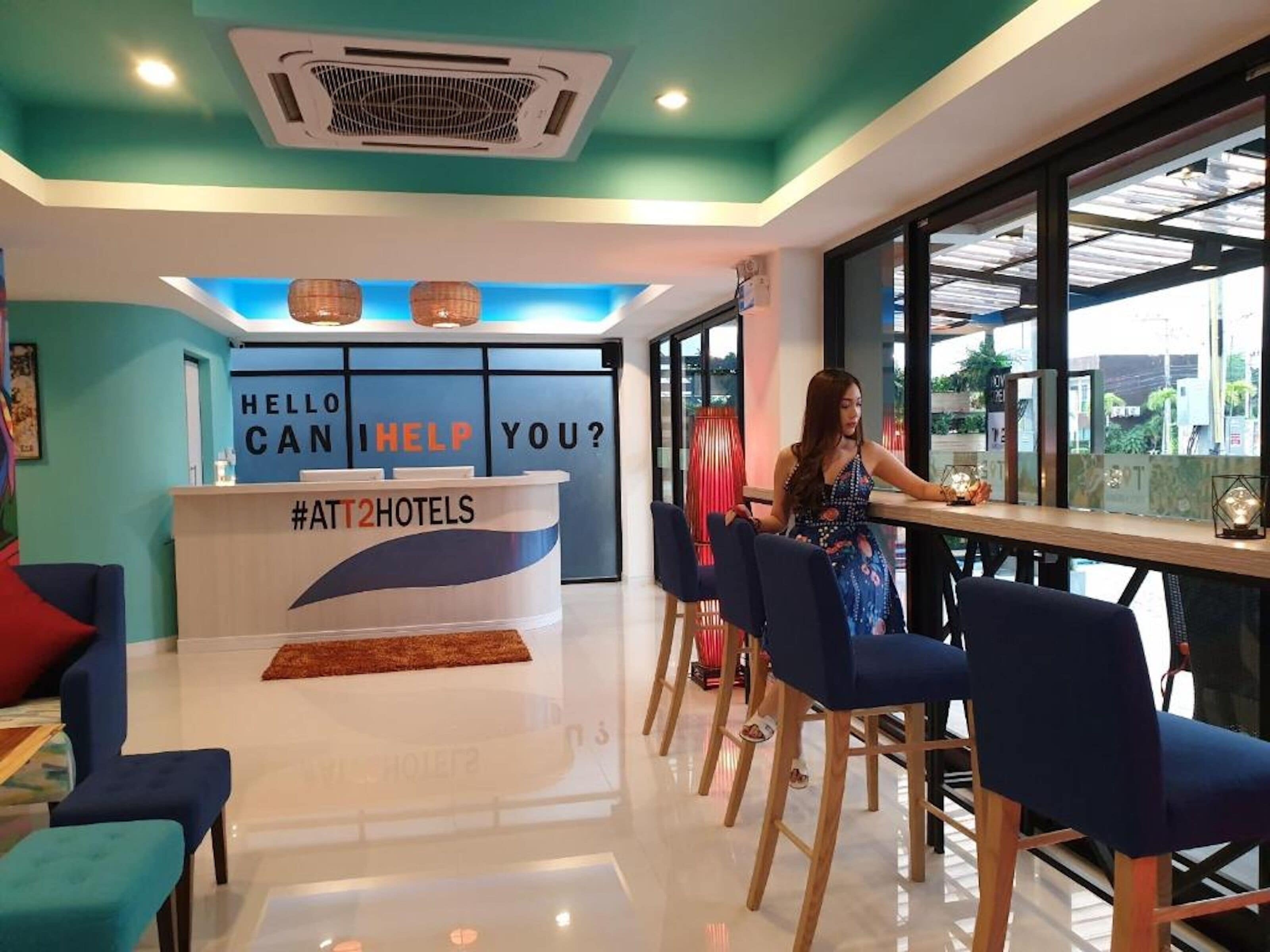 A hotel reception area with a counter displaying "HELLO CAN I HELP YOU?" and a hashtag "#ATT2HOTELS".