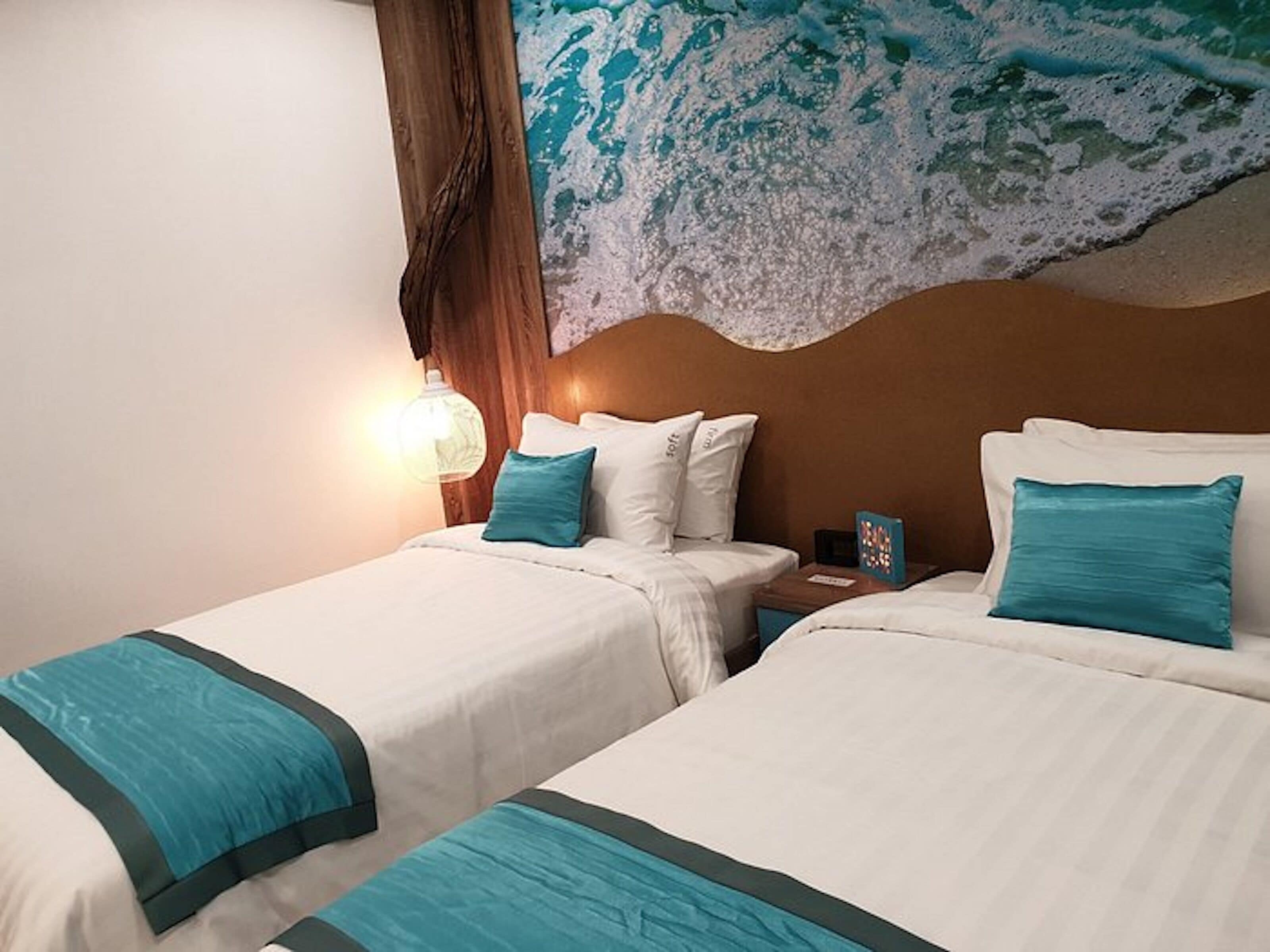 A hotel room with two beds, a large mural of a wave, a wooden headboard, and a nightstand with a lamp.