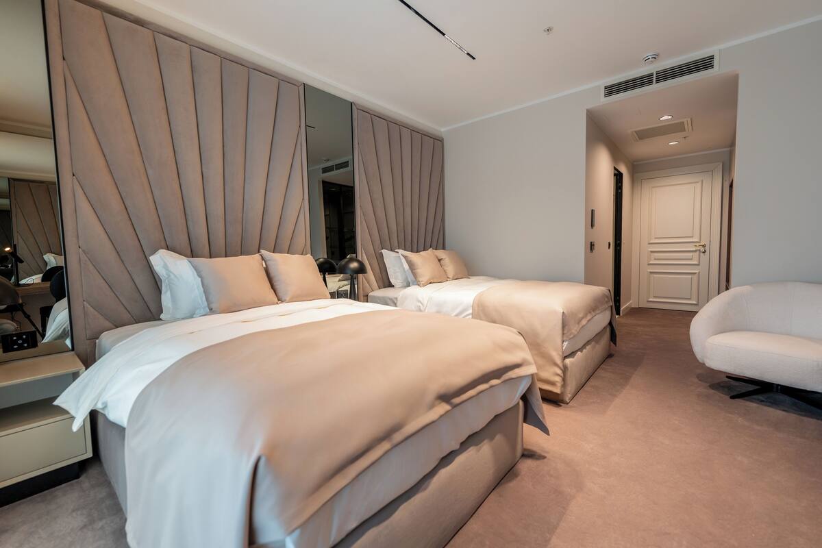 Una habitación de hotel moderna con dos camas, un espejo grande, un sillón blanco y una puerta que da a otra habitación.