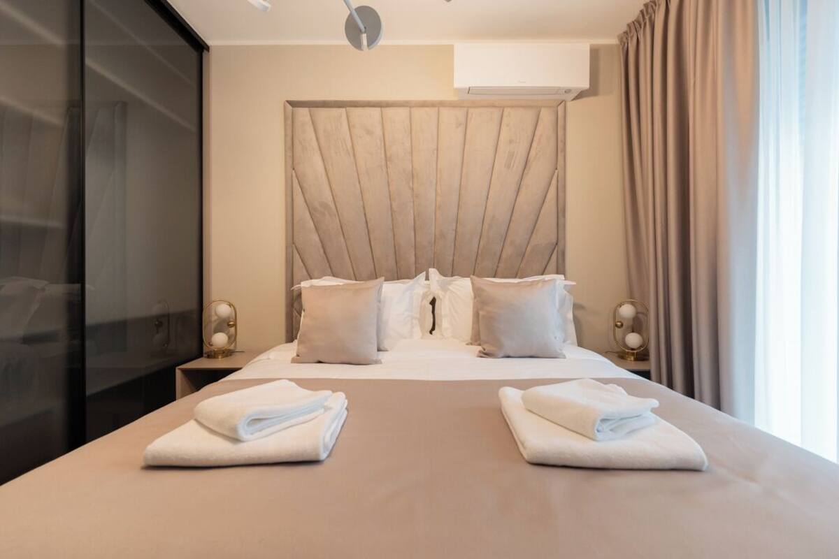 Una habitación de hotel con una cama grande, un cabecero, dos mesitas de noche y una ventana con cortinas.