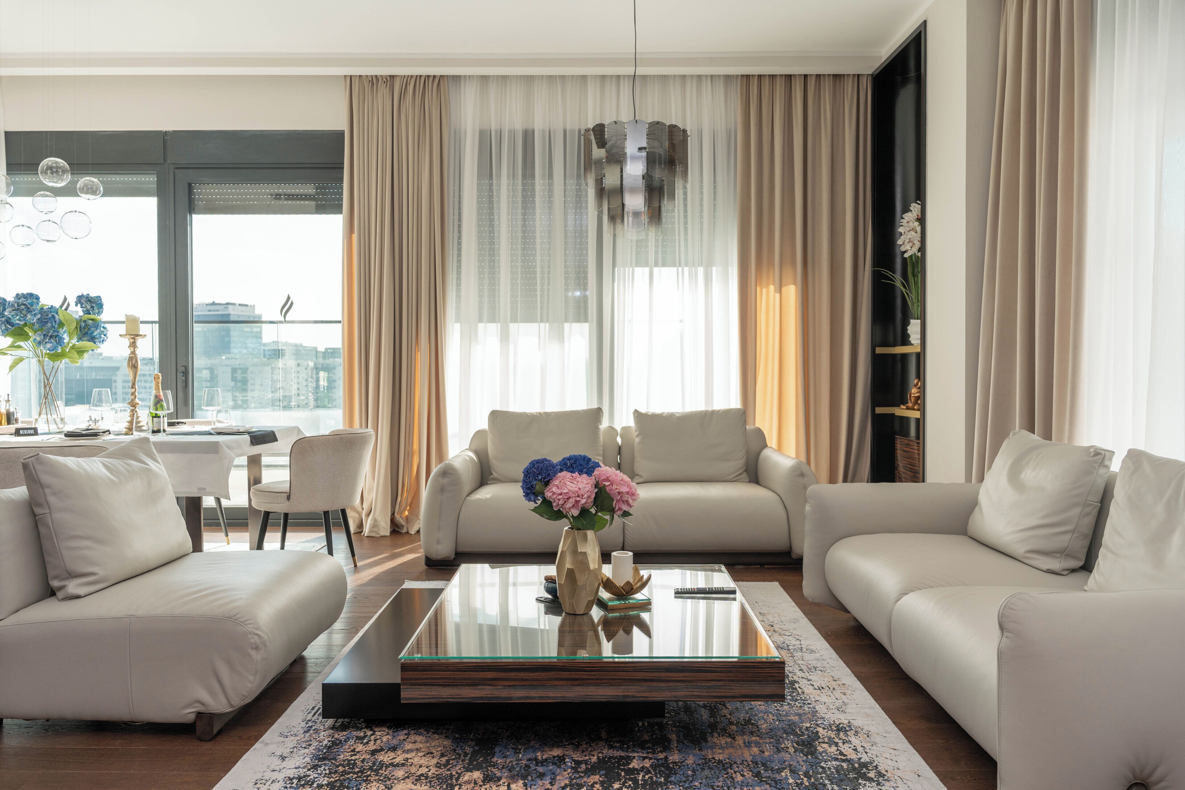 A modern living room with a glass coffee table, two sofas, a dining table, and a view of the city through large windows.