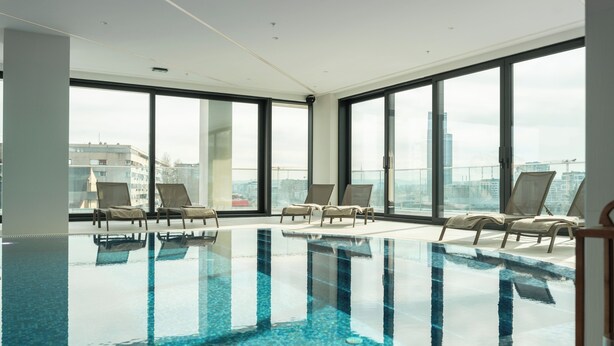 A modern indoor swimming pool with clear glass walls, lounge chairs, and a view of the cityscape. - New Belgrade Premium Hotel (Belgrade)