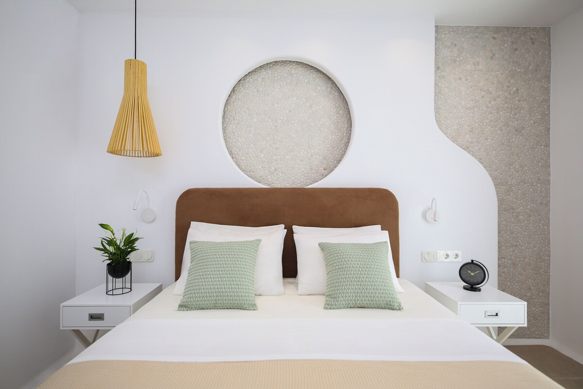 Quarto moderno com cama, duas mesas de cabeceira, luminária circular na parede, relógio de parede e um vaso com planta.