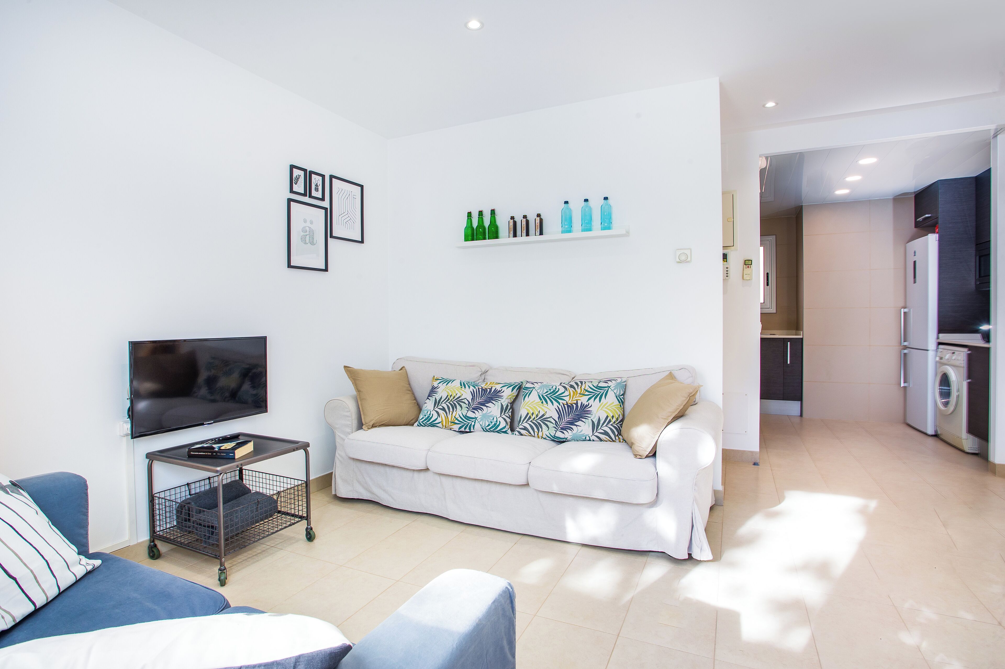 A modern living room with a white sofa, a TV, and a small table. There is a kitchen area visible in the background.