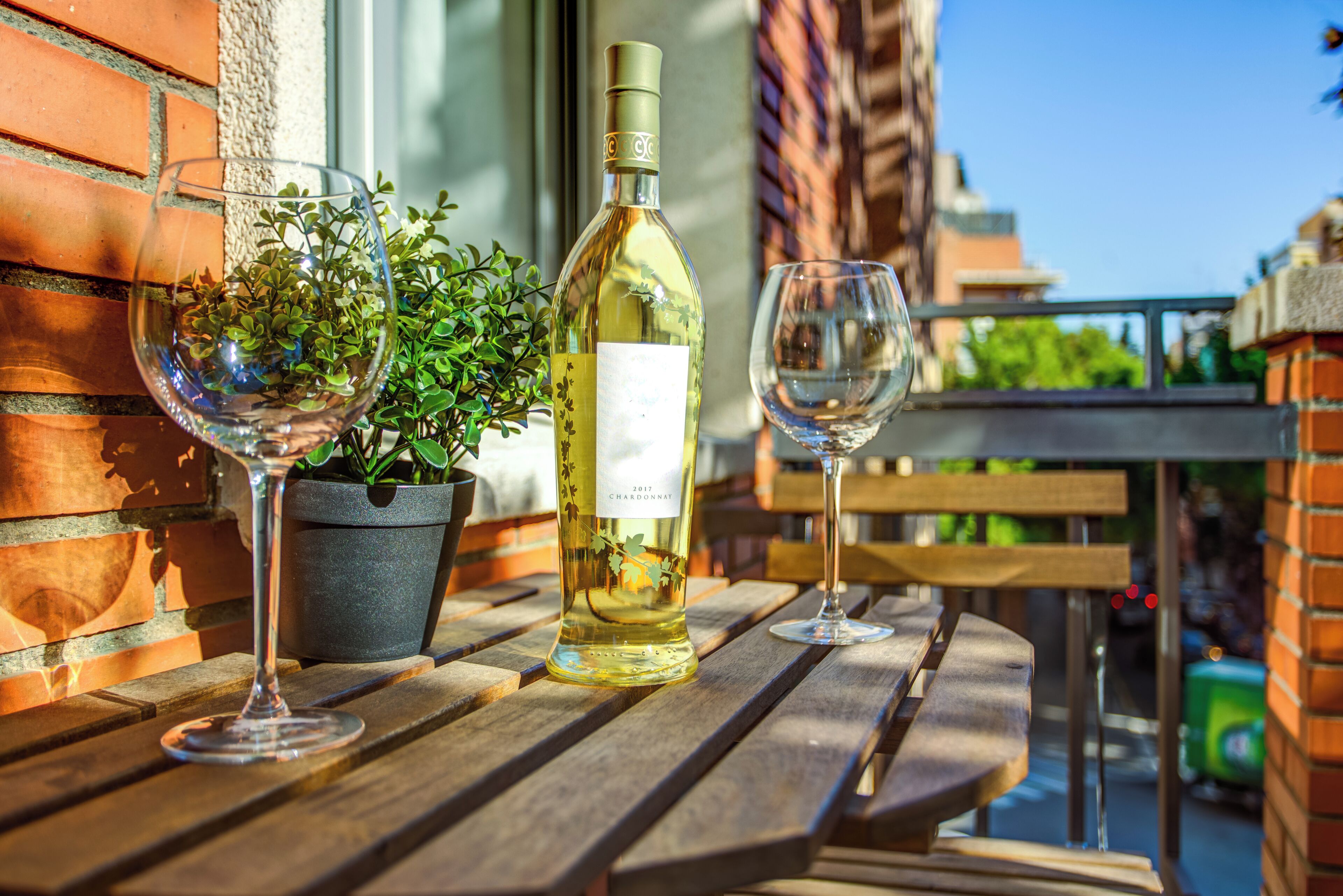 Una botella de vino y dos copas sobre una mesa de madera frente a un edificio con paredes de ladrillo.