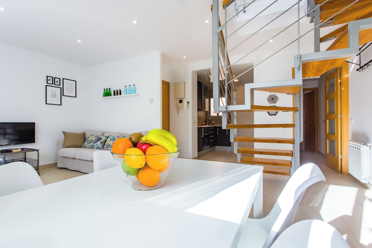 A modern living room with a white sofa, a glass coffee table with fruit, a TV, and a staircase.