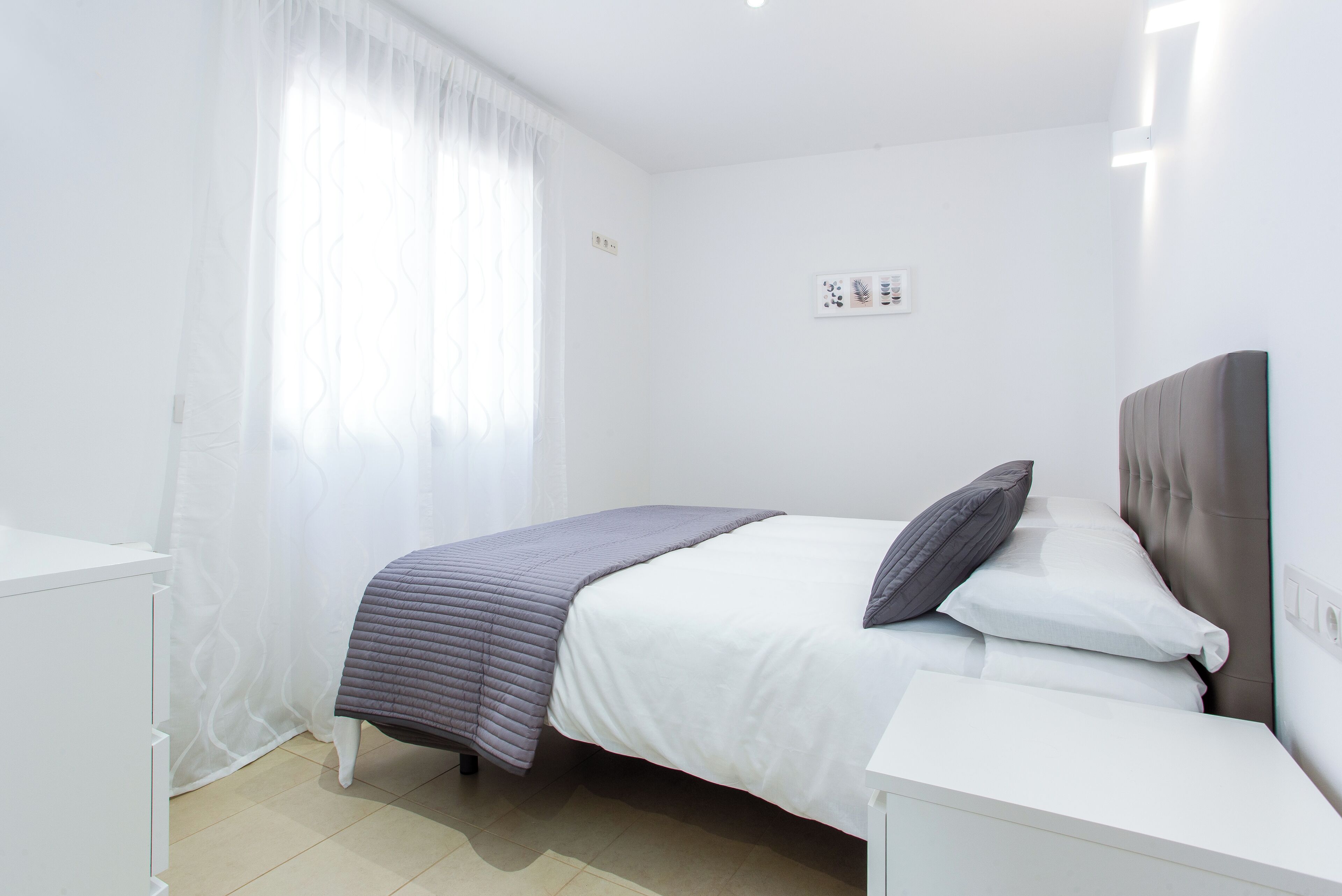 Un dormitorio con cama, paredes blancas, una ventana con cortinas translúcidas y una mesita de noche.