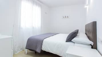 Un dormitorio con cama, paredes blancas, una ventana con cortinas translúcidas y una mesita de noche.