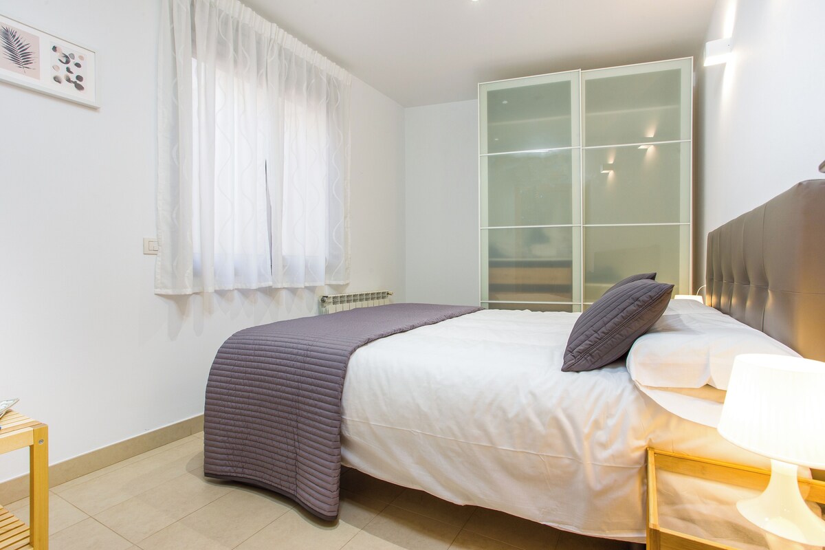 A neatly arranged bedroom with a bed, a bedside table, a lamp, and a window with sheer curtains.