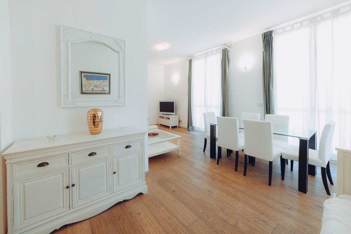 A spacious room with a white dining table and chairs, a TV, and a cabinet with drawers.