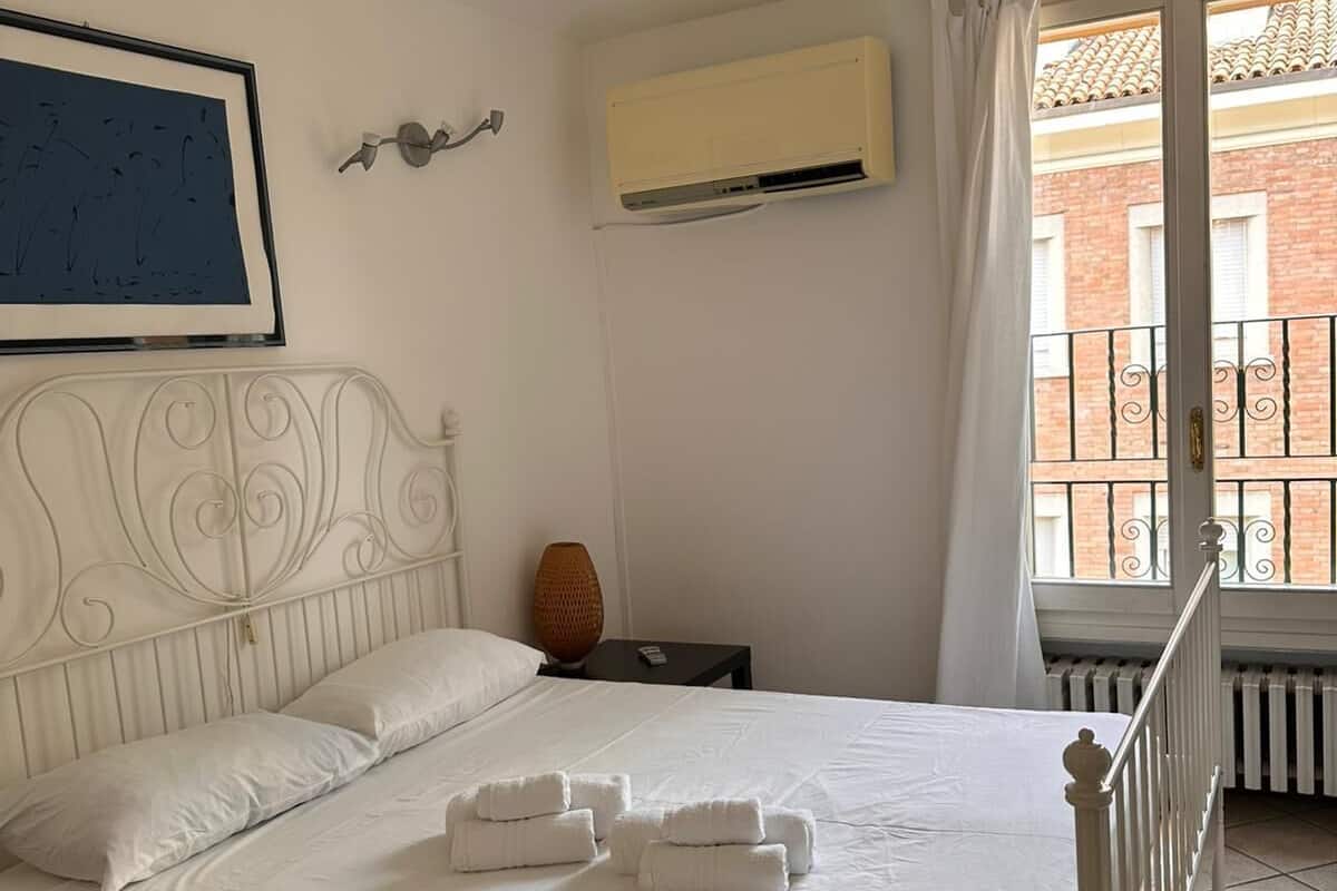 A neatly made bed with white linens and towels, a white headboard, a wall-mounted air conditioner, a framed picture, and a window with a view of a brick building.