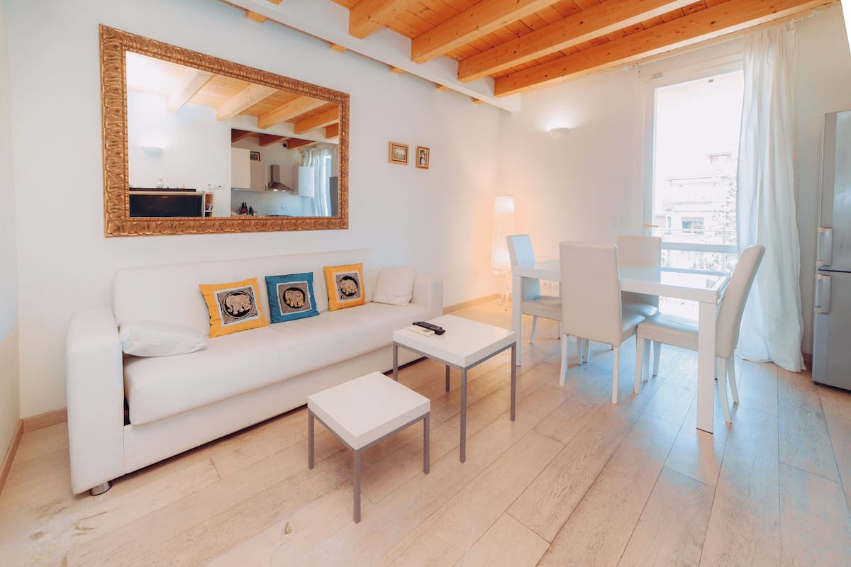 A modern living room with a white sofa, two small tables, a dining area with white chairs, a large mirror, and a kitchen visible through an open door.