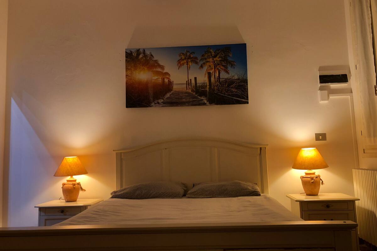 A bedroom with a bed, two bedside tables with lamps, a nightstand, and a painting of a beach scene.