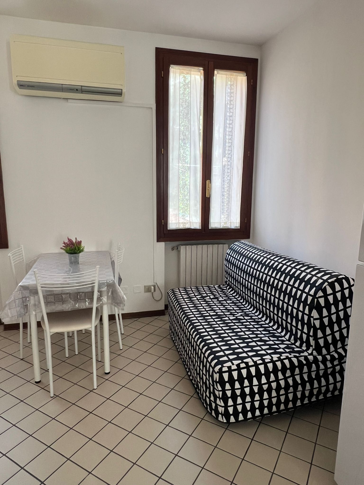 Une petite pièce avec un sol carrelé, une table à manger avec des chaises, un canapé à motifs noir et blanc, une fenêtre avec des rideaux et un climatiseur fixé au mur.