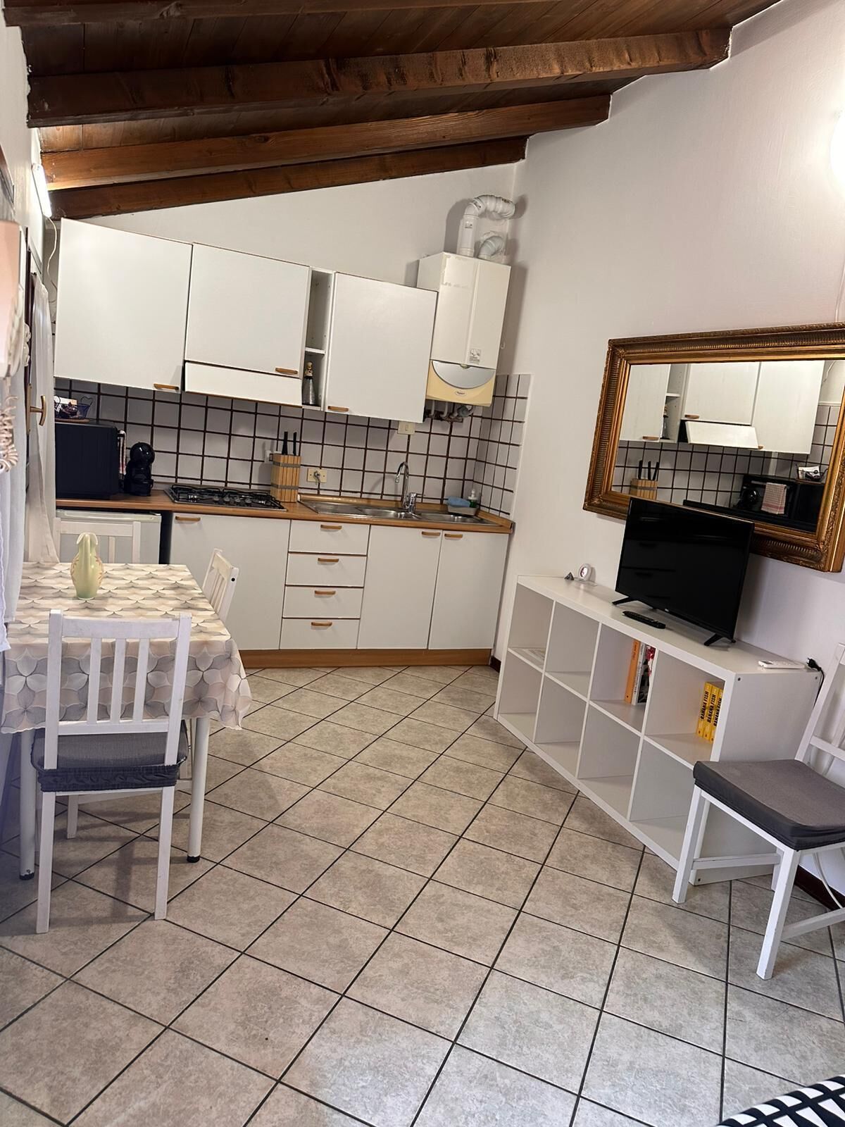 Cozinha compacta com armários brancos, painel de azulejos, teto de madeira, mesa de jantar com cadeiras, TV sobre um suporte branco e uma pequena estante.
