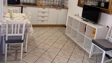 Cozinha compacta com armários brancos, painel de azulejos, teto de madeira, mesa de jantar com cadeiras, TV sobre um suporte branco e uma pequena estante.