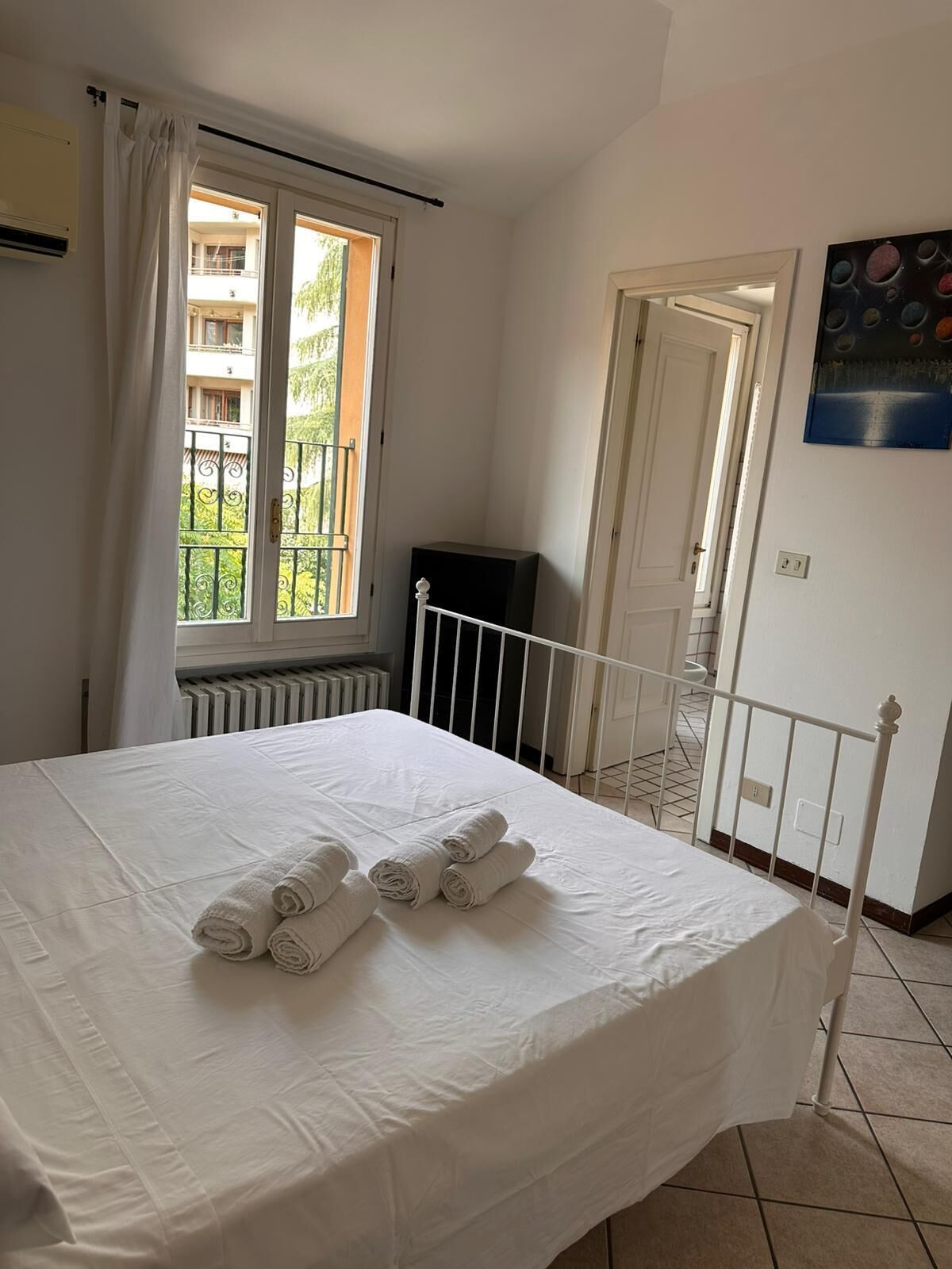 A bedroom with a bed, a radiator, a window with curtains, a door, and a painting on the wall.