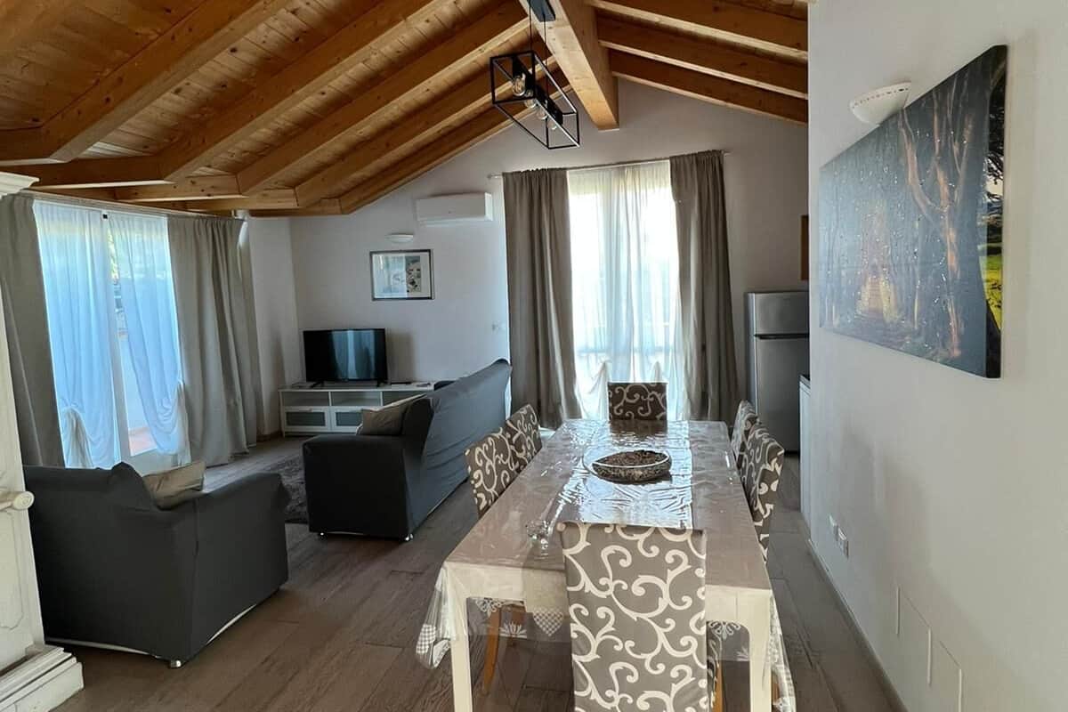 A spacious living area with a high ceiling and wooden beams, a dining table with patterned chairs, a sofa, and a television.