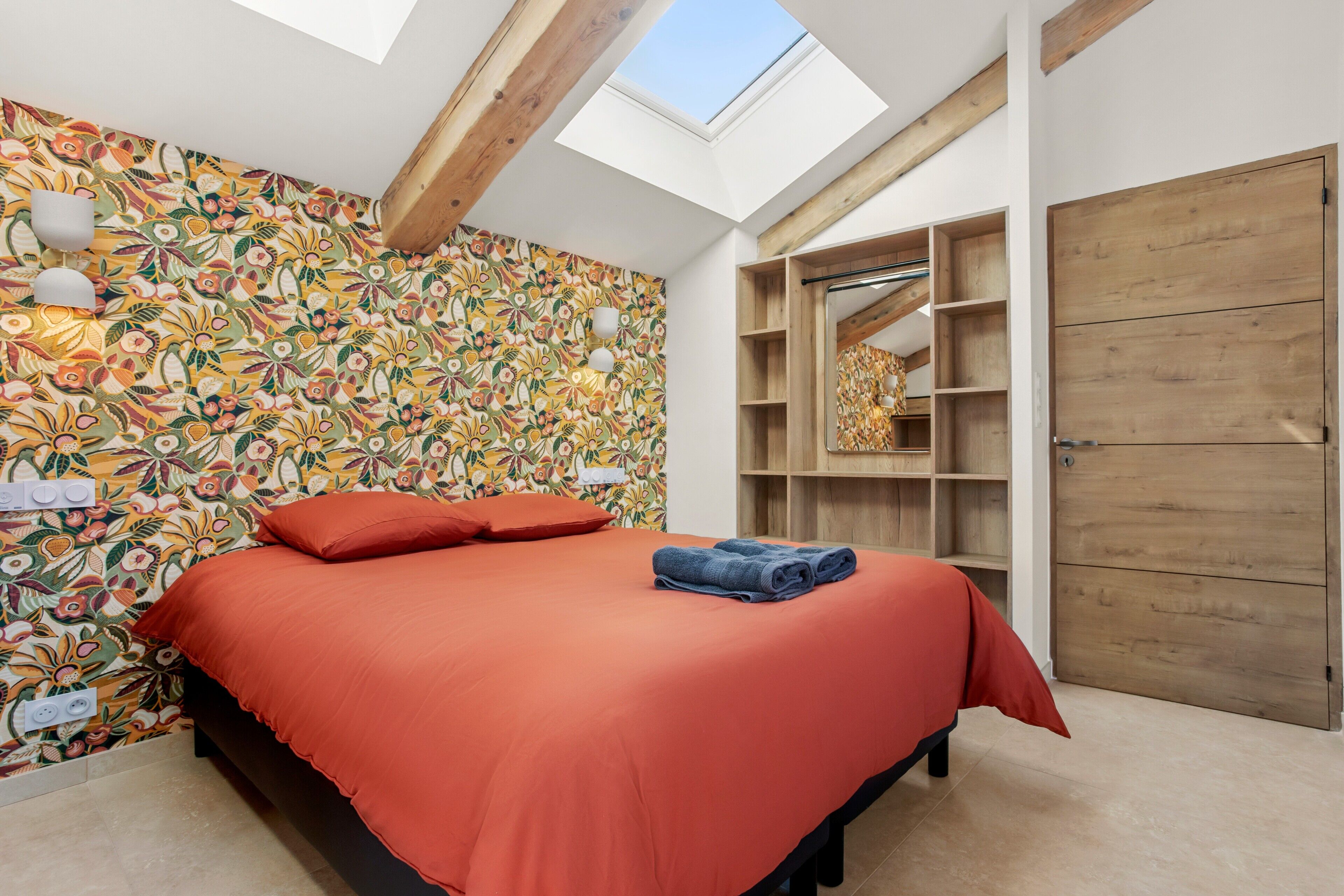 Une chambre dotée d’une lucarne, d’un lit avec une literie rouge, d’un mur à motifs et d’étagères en bois intégrées.